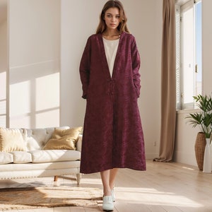 May include: A long, burgundy-coloured coat with a V-neckline and a button closure. The coat has a textured, quilted design and is worn by a person standing in a room with a white sofa and a window.