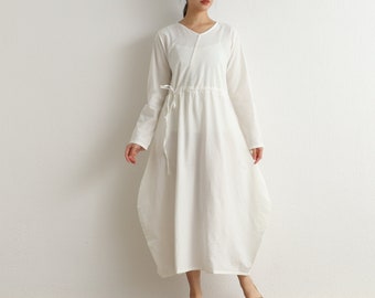 Puede incluir: Un vestido blanco de manga larga con cuello en V y cintura con cordón. El vestido tiene una silueta suelta y fluida.