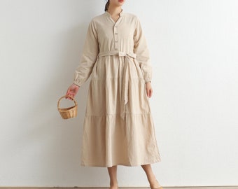 May include: A beige long-sleeve dress with a button-up front, belted waist, and tiered skirt. The dress is paired with yellow flats and a small woven basket. The dress has a mandarin collar.