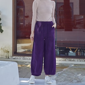 May include: A woman wearing a light brown long-sleeved shirt and wide-leg purple pants. The pants have an elastic waistband and pockets. She is wearing white sneakers. The background includes a window and a white wall.