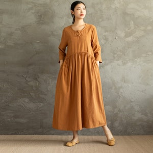May include: A long, loose-fitting, rust-coloured dress with a round neckline and a button closure at the neck. The dress has a gathered waist and long sleeves.