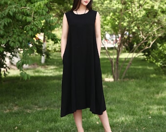 May include: A woman wearing a black sleeveless dress with a simple design. She is standing in a grassy area with trees in the background.