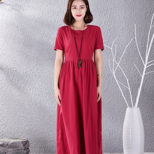 May include: A woman wearing a red linen dress with short sleeves and a round neckline. She is wearing a necklace with a wooden pendant. The dress has a gathered waist and a flowing skirt.