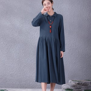 May include: A woman wearing a long-sleeved, blue, collared dress with a red beaded necklace. She is standing in front of a grey textured wall and smiling.