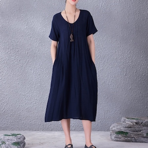 May include: A navy blue, knee-length dress with short sleeves and a V-neck. The dress features pockets and a simple, flowing design. A long necklace with a pendant is worn. The model is wearing dark shoes.