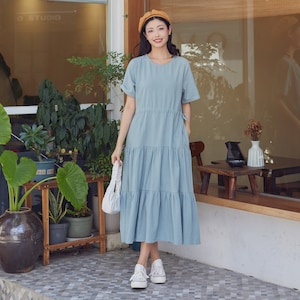 May include: A woman wearing a light blue tiered dress with a brown beret and white sneakers. She is holding a white handbag and standing in front of a building with a window.