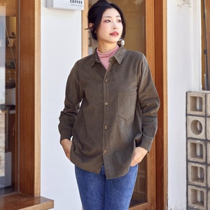 May include: A woman wearing a dark green corduroy shirt with a button-down front and a single pocket on the left side. She is standing in front of a coffee shop with a sign that reads "Coffee Cafe & Store".
