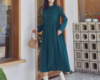 May include: A teal blue long-sleeved dress with a mandarin collar and button closure. The dress has a gathered waist and a slightly flared skirt. The model is wearing white ankle boots and carrying a brown handbag.