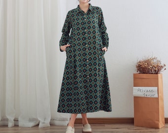 May include: A long-sleeved, green and black patterned dress with a button-up front and pockets. The dress has a slightly flared skirt and is worn by a person with their hands in the pockets. The person is standing in front of a white wall with a brown paper bag that says "Private Tailor" on it.