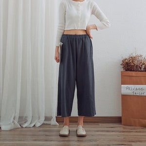 May include: A woman wearing a white cropped cardigan and dark gray wide-leg pants. She is standing in front of a white curtain and a brown paper bag with the text "Private Tailor" on it.