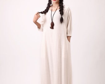 May include: A white linen dress with a scoop neck and 3/4 sleeves. The dress has a loose fit and a gathered hem. The model is wearing a brown wooden pendant necklace.