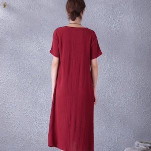 May include: A burgundy-colored midi dress with short sleeves and a scoop neckline. The dress has a textured appearance and falls to mid-calf. The model is wearing black flats. The background is a textured gray wall.