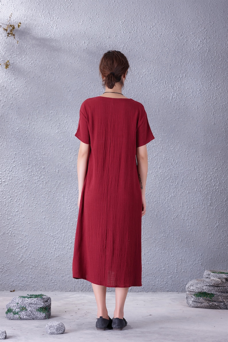 May include: A burgundy-colored midi dress with short sleeves and a scoop neckline. The dress has a textured appearance and falls to mid-calf. The model is wearing black flats. The background is a textured gray wall.