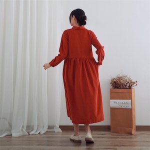 May include: A long-sleeved, red corduroy dress with a collared neckline and a gathered waist. The dress is worn by a person standing in front of a white wall and a sheer white curtain.