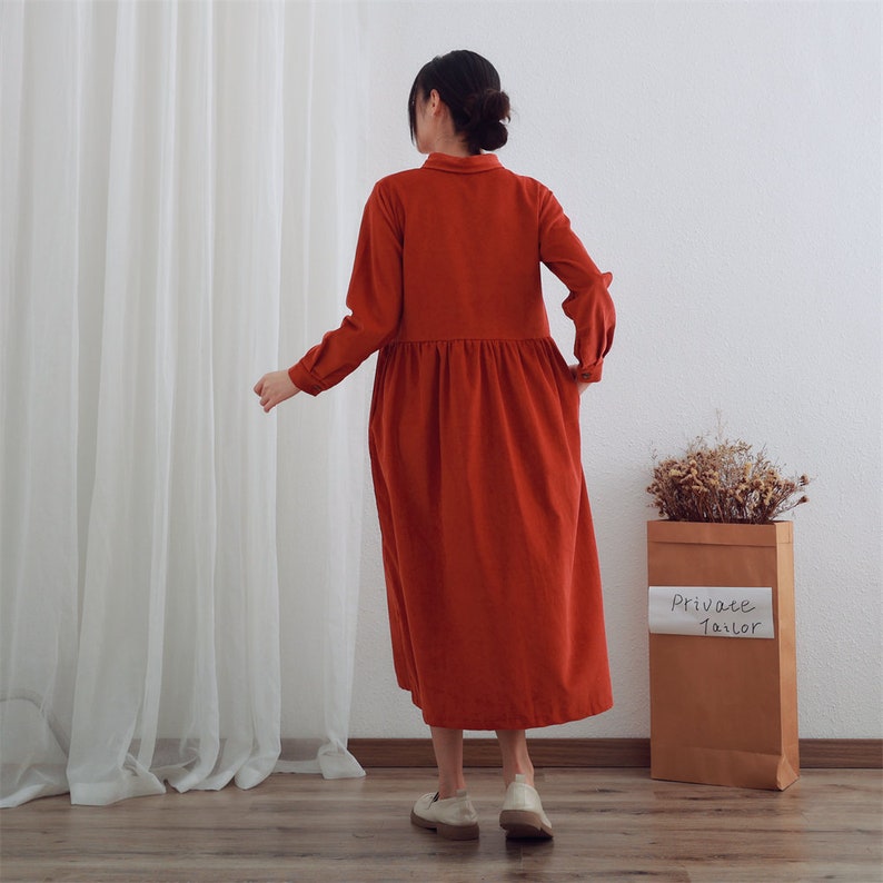 May include: A long-sleeved, red corduroy dress with a collared neckline and a gathered waist. The dress is worn by a person standing in front of a white wall and a sheer white curtain.