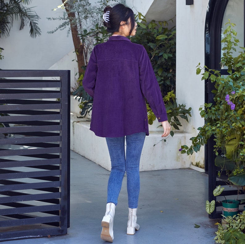 K&ouml;nnte beinhalten: Ein lang&auml;rmeliges, lila Cordhemd mit Knopfleiste und Seitenschlitzen. Das Hemd wird mit einer blauen Jeans und wei&szlig;en Stiefeletten getragen. Das Outfit wird im Freien getragen.