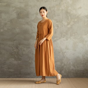 May include: A long, loose-fitting, rust-colored dress with a mandarin collar and a button closure at the neckline. The dress has 3/4 sleeves and a gathered skirt.