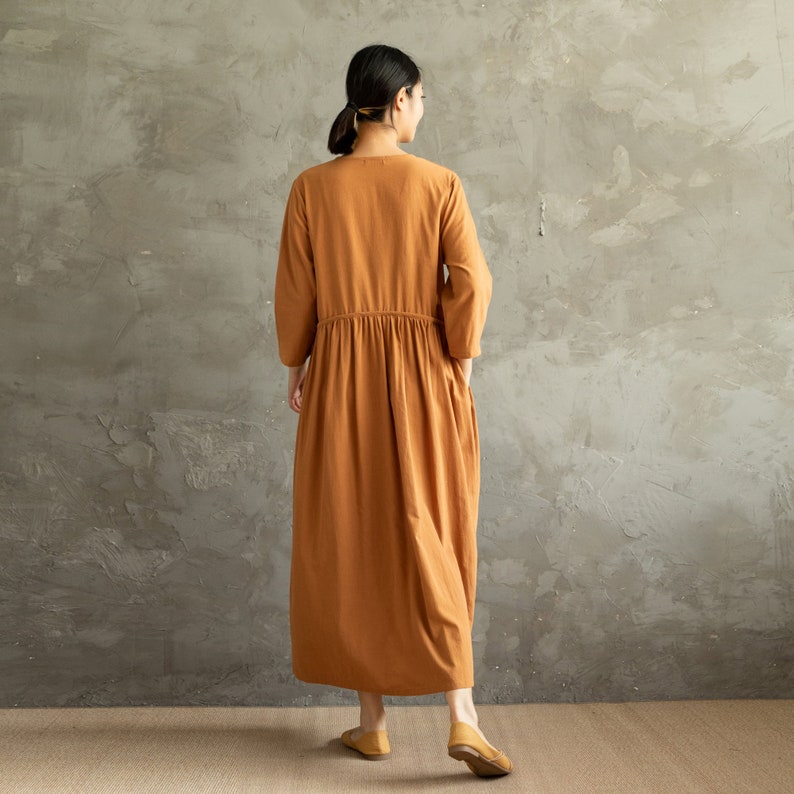 May include: A long, rust-colored dress with a gathered waist and long sleeves. The dress is made of a lightweight fabric and has a relaxed fit.