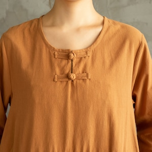 May include: A close-up of a brown linen shirt with a round neckline and a button closure at the neck. The shirt has long sleeves and a relaxed fit.