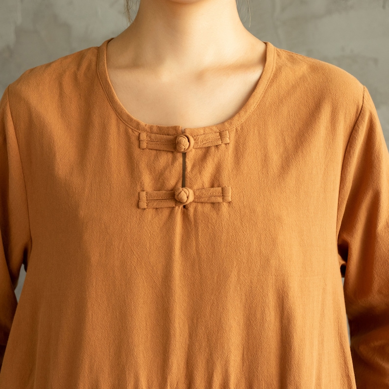 May include: A close-up of a brown linen shirt with a round neckline and a button closure at the neck. The shirt has long sleeves and a relaxed fit.