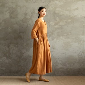May include: A long, loose-fitting, rust-colored dress with a round neckline and three-quarter sleeves. The dress has a gathered waist and pockets on the sides.
