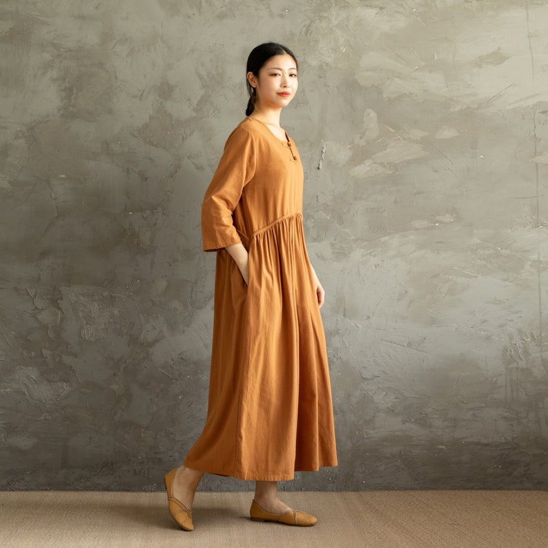 May include: A long, loose-fitting, rust-colored dress with a round neckline and three-quarter sleeves. The dress has a gathered waist and pockets on the sides.