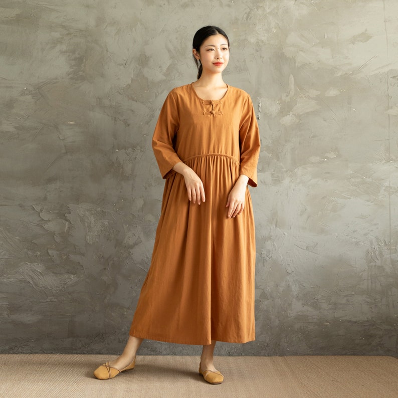 May include: A long, loose-fitting, rust-colored dress with a round neckline and a button closure at the neck. The dress has a gathered waist and long sleeves.