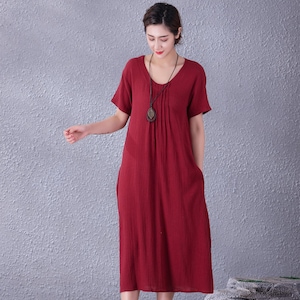 May include: A midi-length, short-sleeved dress in a deep red hue. The dress features a scoop neckline and a pleated bodice detail. It has pockets and is made of a textured fabric. A long necklace with a pendant is worn with the dress.