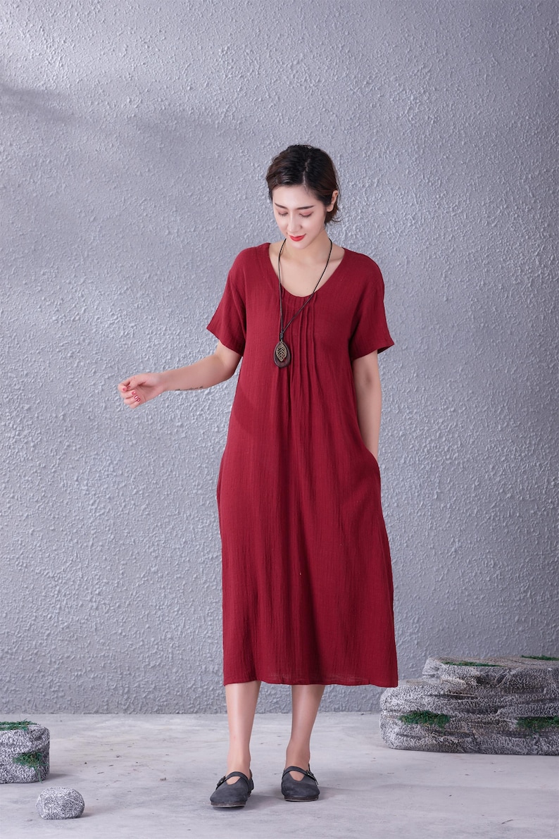May include: A midi-length, short-sleeved dress in a deep red hue. The dress features a scoop neckline and a pleated bodice detail. It has pockets and is made of a textured fabric. A long necklace with a pendant is worn with the dress.