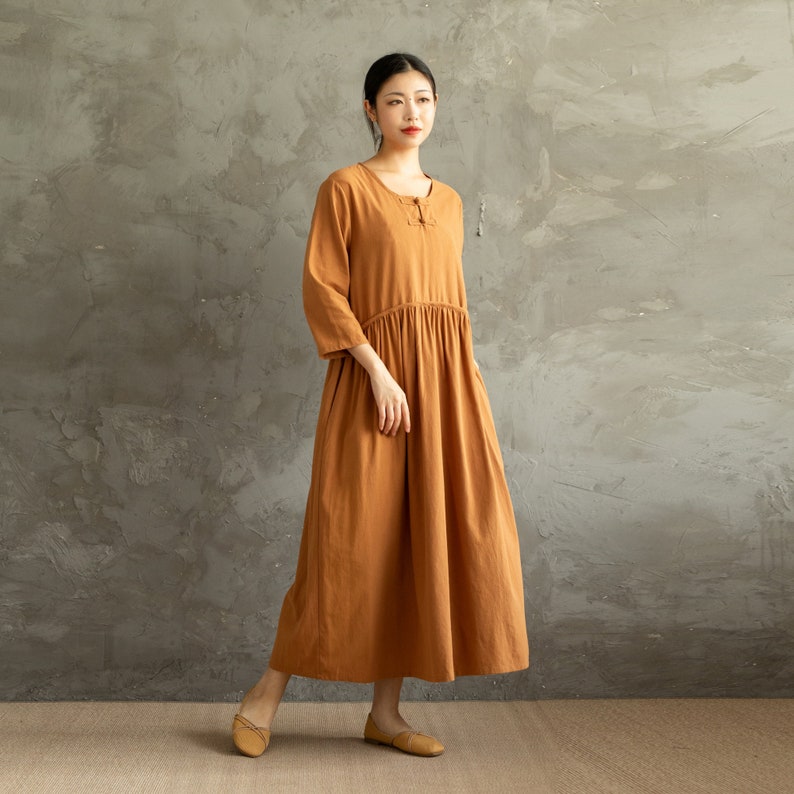 May include: A long, loose-fitting, rust-colored dress with a round neckline and three-quarter sleeves. The dress has a gathered waist and a simple, casual style.