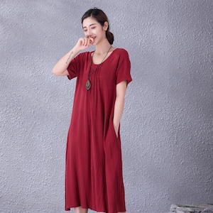 May include: A red midi dress with short sleeves and a round neckline. The dress has a relaxed fit, pockets, and a pleated detail at the neckline. A long necklace with a pendant is worn with the dress. The model is wearing dark shoes.