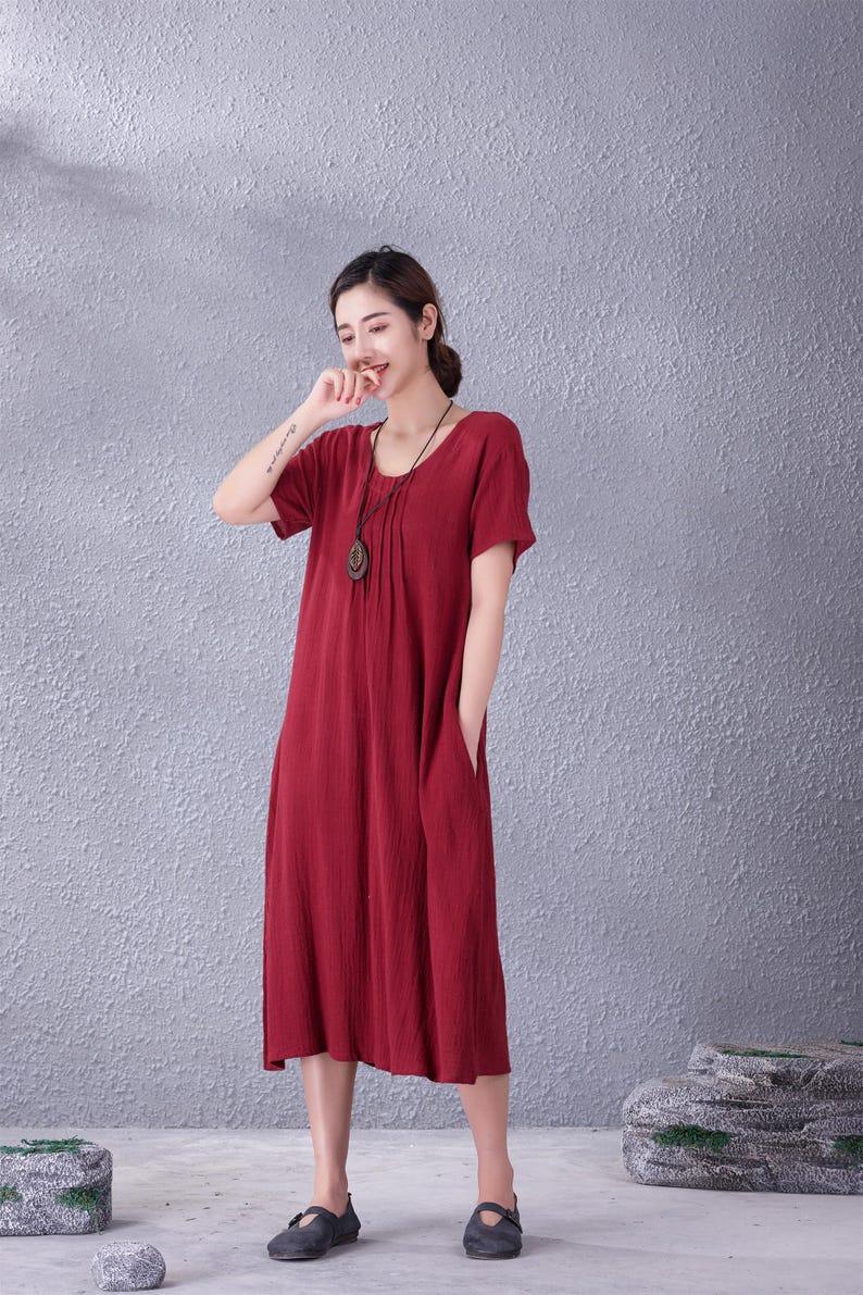 May include: A red midi dress with short sleeves and a round neckline. The dress has a relaxed fit, pockets, and a pleated detail at the neckline. A long necklace with a pendant is worn with the dress. The model is wearing dark shoes.