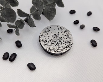 May include: A silver-toned, round compact mirror with a detailed engraved design featuring a fairy seated at a mushroom table, surrounded by leaves and other whimsical elements. Black beans and greenery are scattered around the mirror on a white surface.