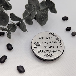 May include: A round, silver-colored metal object with the phrase "Do you suppose she's a wildflower?" etched on it, surrounded by floral and leaf designs. Black beans and greenery are scattered around the object.