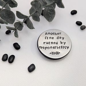 May include: A silver-colored round metal object with the text "Another fine day ruined by responsibility" stamped on it. The object is surrounded by black beans and a sprig of green leaves.