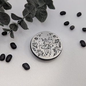 May include: A silver-colored, round compact mirror with a black rim. The mirror's surface is etched with a floral and fairy design. Black beans and a sprig of green leaves are scattered around the mirror on a white surface.