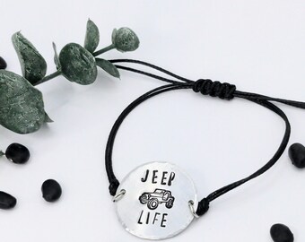 May include: A silver-colored bracelet with a black cord. The bracelet features a circular charm with the words "JEEP LIFE" and a Jeep vehicle illustration. The background is white with a few black beans and a green plant.
