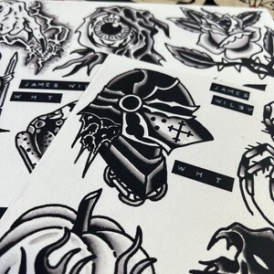 May include: Black and white tattoo flash art sheets featuring traditional designs. The designs include a knight's helmet, roses, an eye, and other gothic-inspired illustrations. The artist's name, James Wilby, is printed on the sheets.