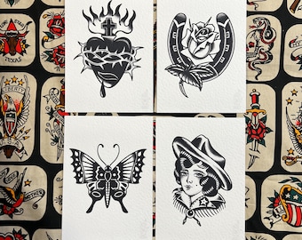 Op de afbeelding: Vier zwart-wit tattoo kunstprints. De prints tonen een hart met een kruis, een hoefijzer met een roos, een vlinder en een vrouw met een hoed. De achtergrond is een stof met diverse tattoo-thema ontwerpen.