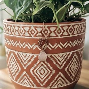 May include: A terracotta-coloured ceramic plant pot with a white geometric pattern. A copper-coloured wire plant stake with a spiral design and a teardrop-shaped rose quartz crystal hangs from the pot, with a green plant inside.