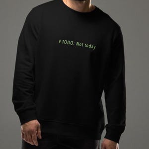 May include: Black crew neck sweatshirt with the text "#TODO: Not today" in green. The sweatshirt is a solid black color and has a relaxed fit. The text is in a simple sans-serif font.