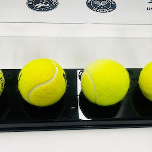 Könnte beinhalten: Vier gelbe Tennisbälle mit weißen Nähten werden auf einem schwarzen Sockel in einer transparenten Vitrine präsentiert. Logos großer Tennisturniere sind darüber zu sehen.