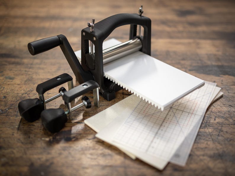 May include: A black metal roller press with a handle and two clamps. The press is on a wooden surface with a white sheet of paper and a gridded sheet of paper.