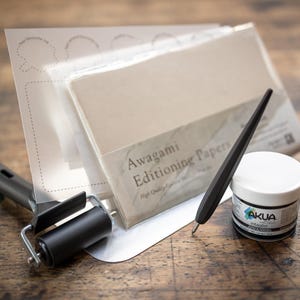 May include: A set of paper crafting tools including a black roller, a black stylus, a small jar of black ink, and a stack of Awagami Editioning Papers. The papers are white and have a label that reads "Awagami Editioning Papers High Quality"