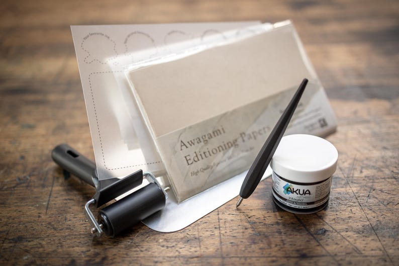 May include: A set of paper crafting tools including a black roller, a black stylus, a small jar of black ink, and a stack of Awagami Editioning Papers. The papers are white and have a label that reads "Awagami Editioning Papers High Quality"