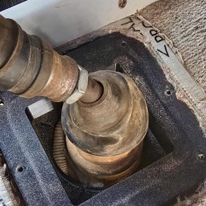 May include: Close-up of a black metal gear shift boot with a brown leather gear shift knob. The boot is secured with screws and is surrounded by a gray carpet.