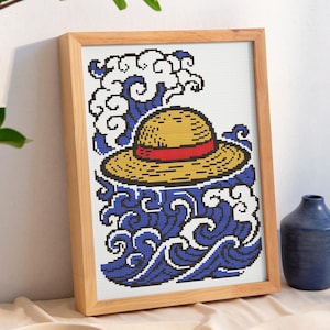 Peut inclure: Une œuvre d'art au point de croix encadrée représentant un chapeau de paille jaune avec une bande rouge sur des vagues bleues. L'œuvre est dans un cadre en bois clair. Un vase bleu et de petites assiettes sont également dans l'image.