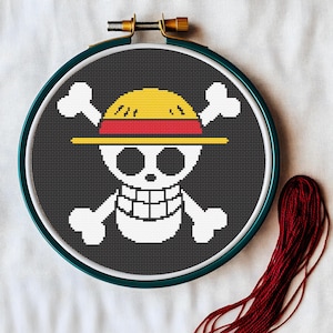 May include: A cross-stitch embroidery hoop featuring a skull and crossbones design with a yellow straw hat and red band. The design is set against a black background, with white cross-stitch details. A bundle of red thread is visible.