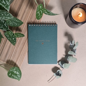Könnte beinhalten: Ein blaugrünes Notizbuch mit einer goldenen Spiralbindung und dem Text "Live joyfully" mit einem goldenen Herzsymbol. Das Notizbuch liegt auf einer braunen Oberfläche mit einer grünen Pflanze und einer brennenden Kerze.