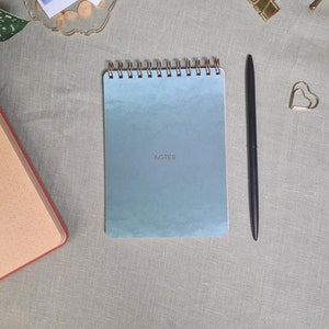 Könnte beinhalten: Ein blaues Spiralnotizbuch mit dem Wort "NOTES" auf dem Einband. Ein schwarzer Stift liegt neben dem Notizbuch auf einer hellgrauen Leinenoberfläche.